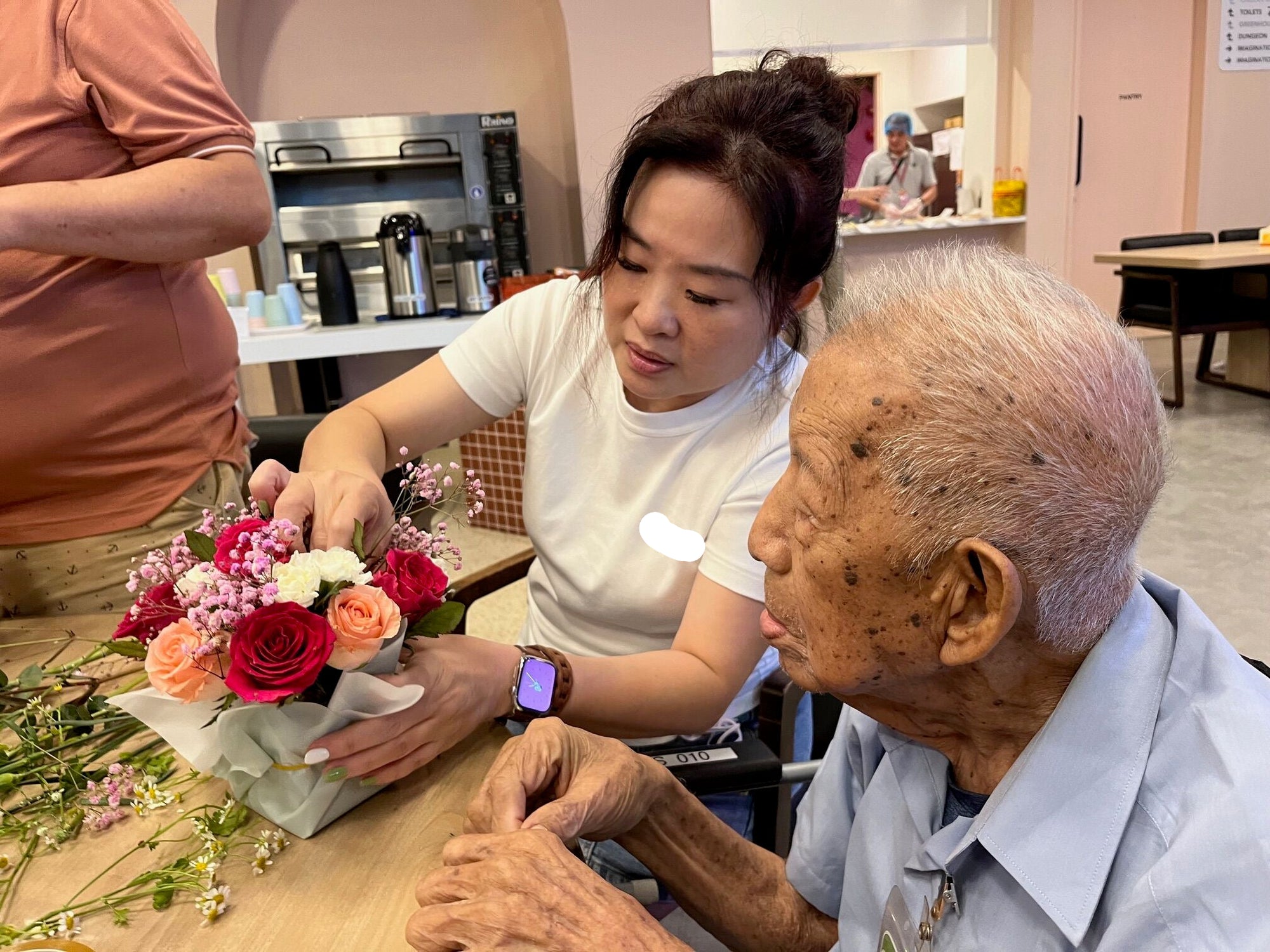 Woman assisting an elderly man with a flower arrangement in a kitchen setting