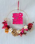 A circular hanging wreath decoration with preserved flowers, in shades of pink, gold, and multicolor, with a red hanging ribbon.