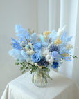 A blue bouquet of dried and preserved flowers arranged in a clear jar, with a variety of flowers including roses and hydrangeas, displayed against a neutral background.