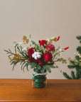 Bouquet of red, white, and gold flowers in a green vase on a wooden surface with a Christmas tree in the background.