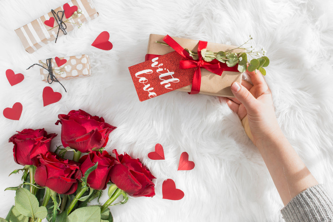 a hand holding a valentines gift and 6 roses on top a fur table cover