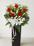 A large floral arrangement in a tall black box, featuring red and white flowers with green foliage and a large white bow, photographed against a plain light gray wall and floor.