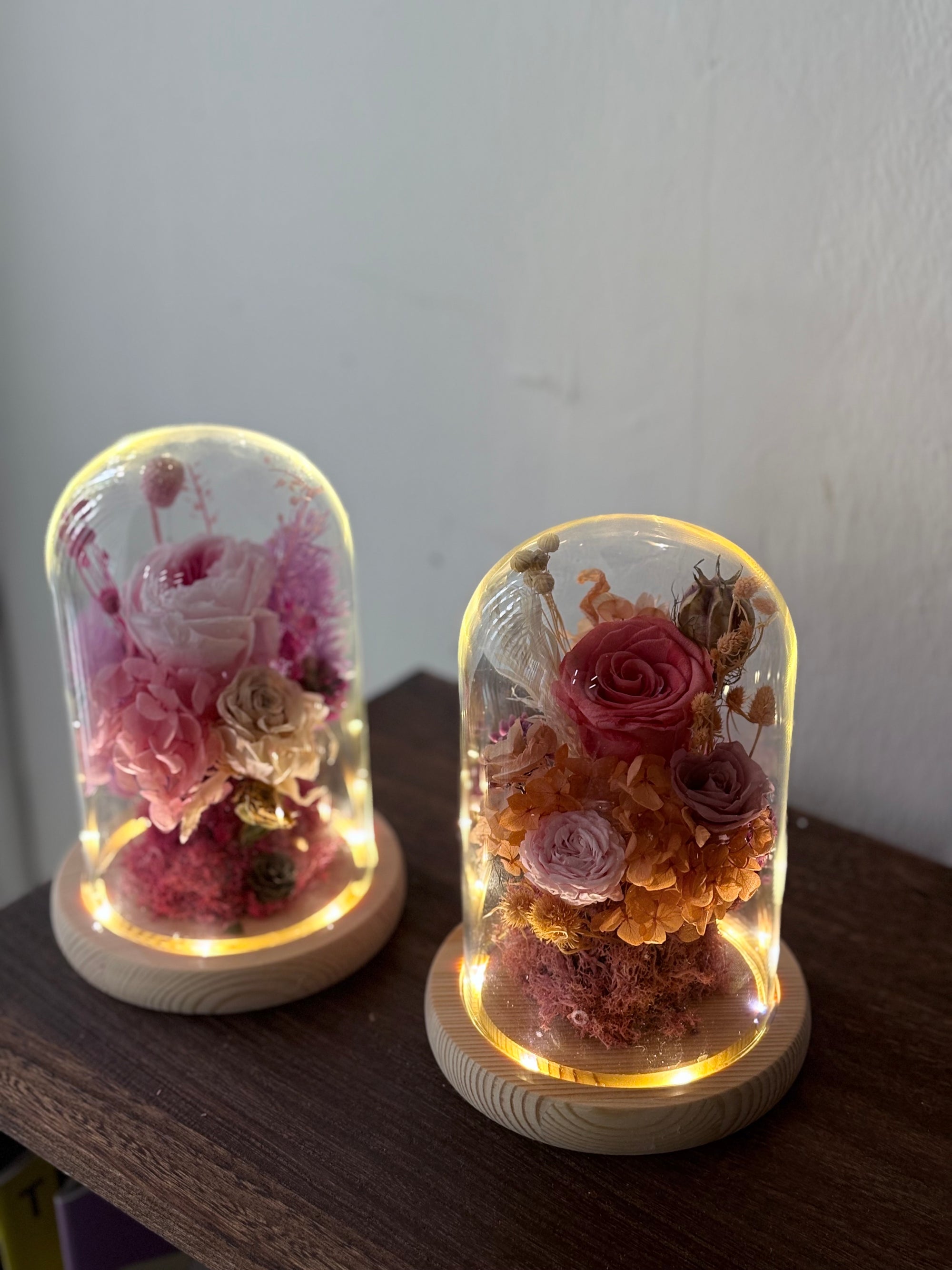 Two illuminated glass dome flower keepsakes on wooden bases, displayed on a dark wooden surface against a plain light wall.