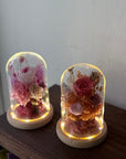 Two illuminated glass dome flower keepsakes on wooden bases, displayed on a dark wooden surface against a plain light wall.