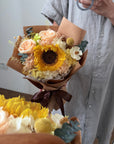 Person holding a bouquet of flowers with sunflowers and other flowers, wearing a light gray shirt.