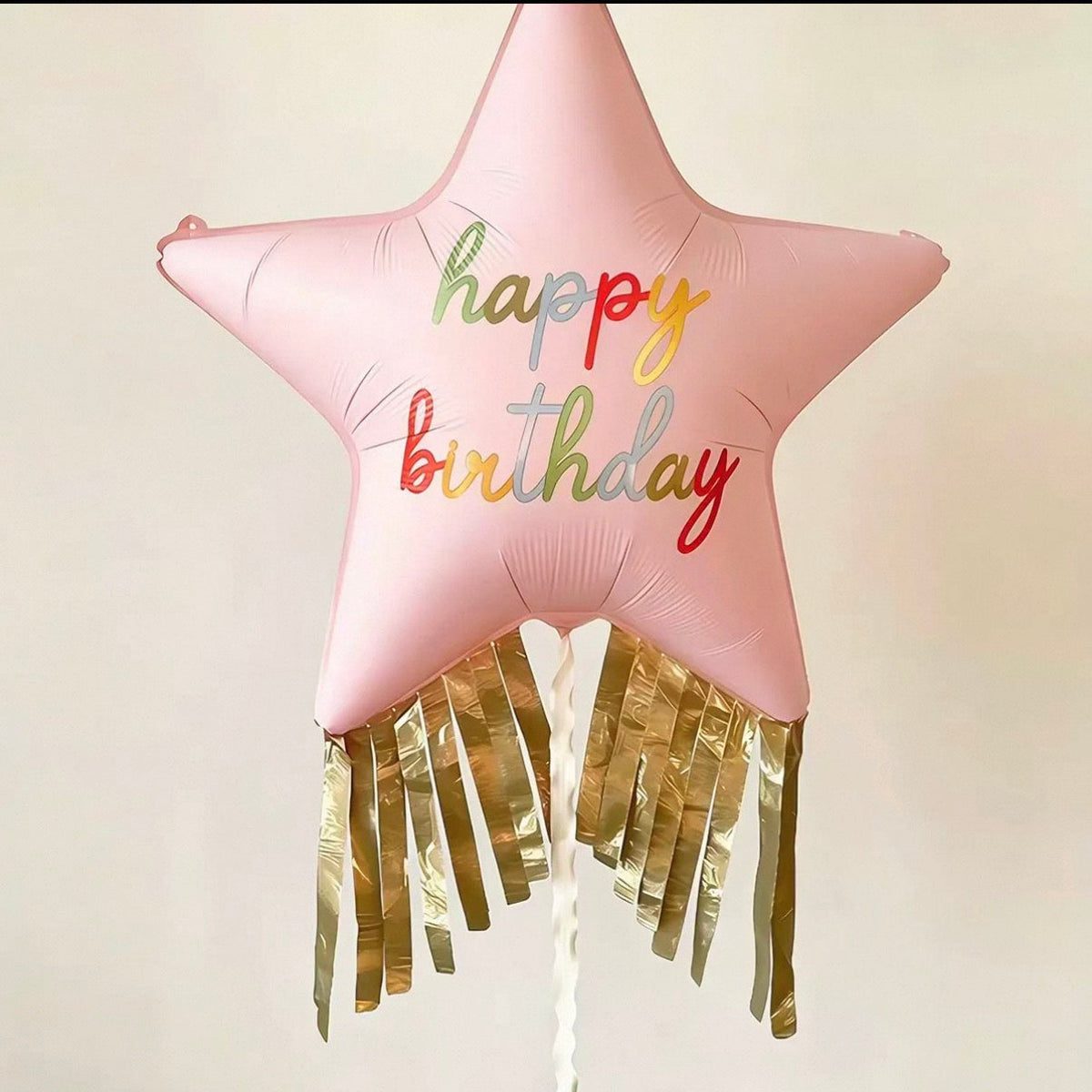 A pink star-shaped foil balloon with 'happy birthday' written in colorful script, finished with gold tinsel fringe at the bottom, shown against a plain light background.