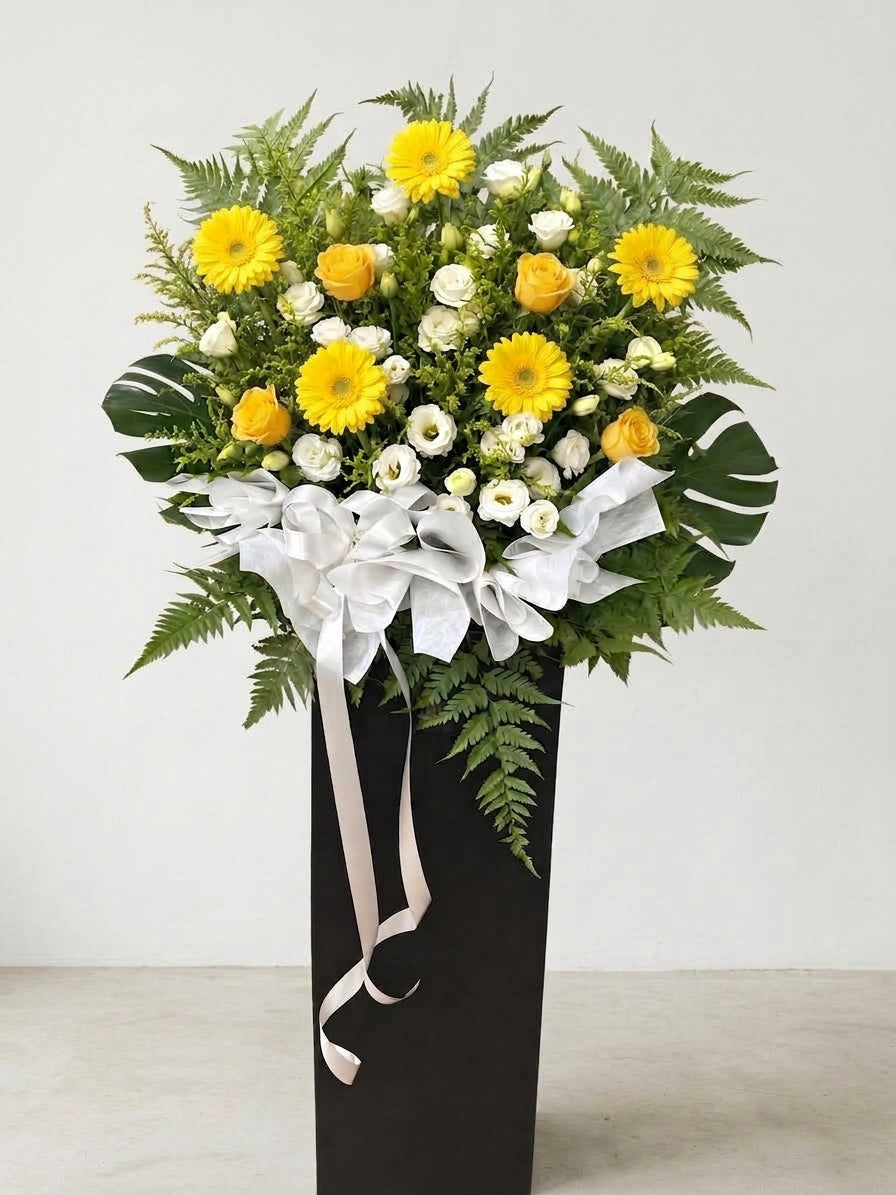 A condolences floral arrangement featuring yellow gerbera daisies, yellow roses, white carnations, and green ferns, presented in a tall black box with a large white bow and ribbon.
