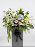 A large floral arrangement featuring light pink roses, blue hydrangeas, white lilies, and green foliage, presented in a tall gray box tied with a black ribbon, against a plain light gray background.