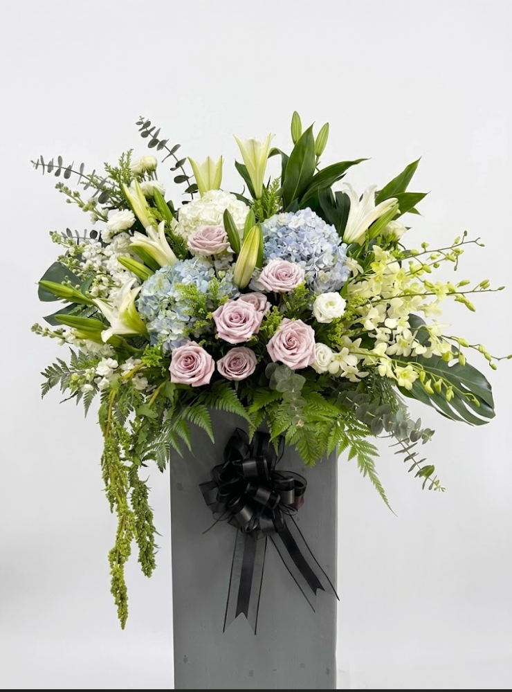 A large floral arrangement featuring light pink roses, blue hydrangeas, white lilies, and green foliage, presented in a tall gray box tied with a black ribbon, against a plain light gray background.