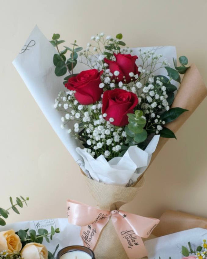 Bouquet of red roses with baby's breath, wrapped in white paper with a pink bow, on a beige background.