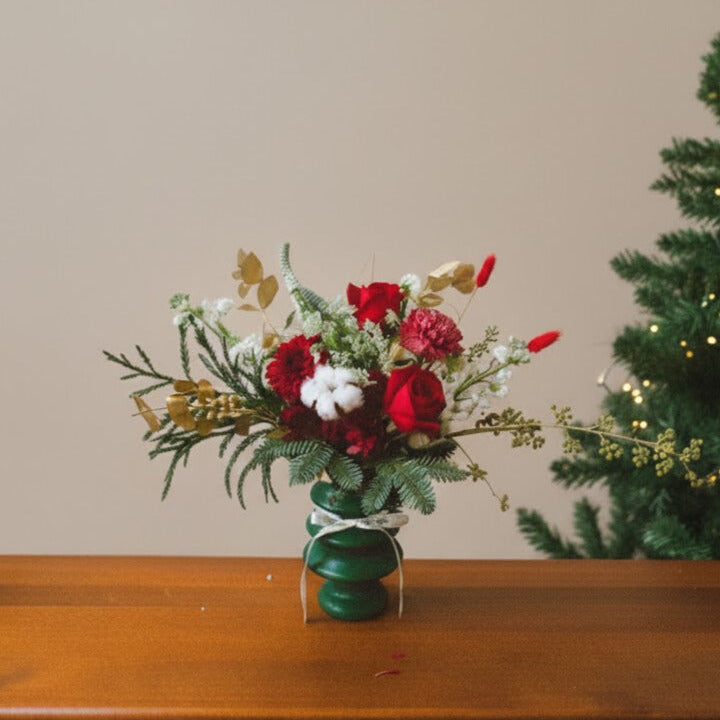 Bouquet of red, white, and gold flowers in a green vase on a wooden surface with a Christmas tree in the background.